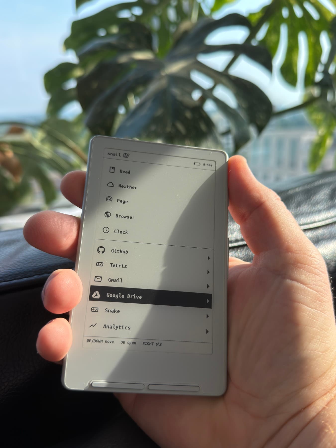 An XTEINK X4 running Snail OS, held in a hand in front of a window, the launcher on its screen.